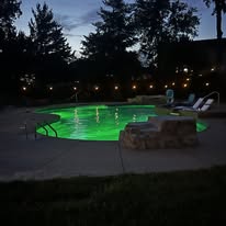 Pool area at night with green water, lounge chairs, and a small table.