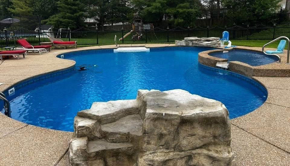 Swimming pool with rock-like structure and lounge chairs in a backyard setting.